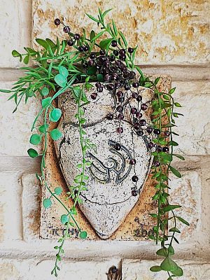 עציץ קיר דקורטיבי עשוי חומר קרמי ממנו משתרכים צמחים ירוקים וסגולים. הפריט מעוצה בסגנון כפרי בעל 