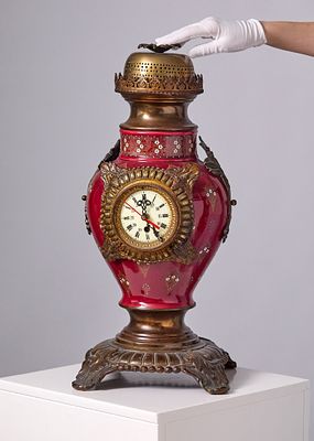שעון שולחני מרשים בעל גוף חרסינה צבועה באמייל בגוון אדום עמוק. משולב בעיטורי נחושת ופליז עם לוח 
