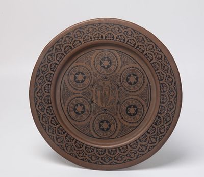 צלחת פסח לתלייה, ארה"ב ראשית שנות ה-20, עשויה פליז, במרכז רשום פסח, זרוע, חרוסת, חזרת, כרפס 