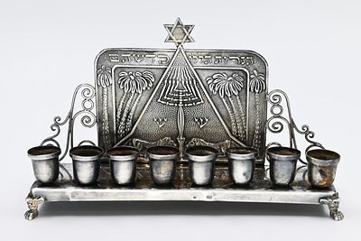 חנוכייה כסף, ארץ ישראל, ראשית שנות ה-20. חתום. 
בבסיס הדופן האחורית ניצבים שני אריות התומכים 