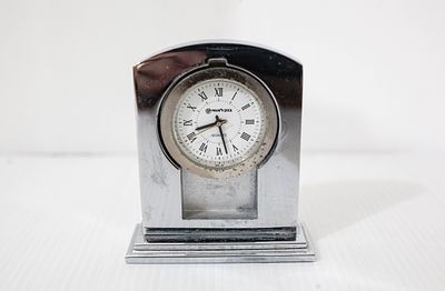שעון שולחני קטן בעל גוף מתכת כסוף ומבריק, הכולל לוח שעון לבן עם ספרות רומיות ואת לוגו "בנק 