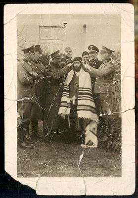 תצלום מתקופת השואה ובו חיילים גרמנים גוזזים את פאותיו של יהודי חרדי העטוף בטלית, בעוד הניצבים 