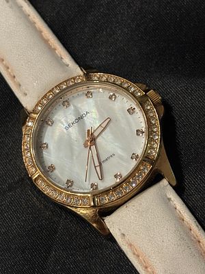 שעון SEKONDA בעיצוב אלגנטי עם רצועת עור לבנה ופנים מכתשית מוקף באבני חן