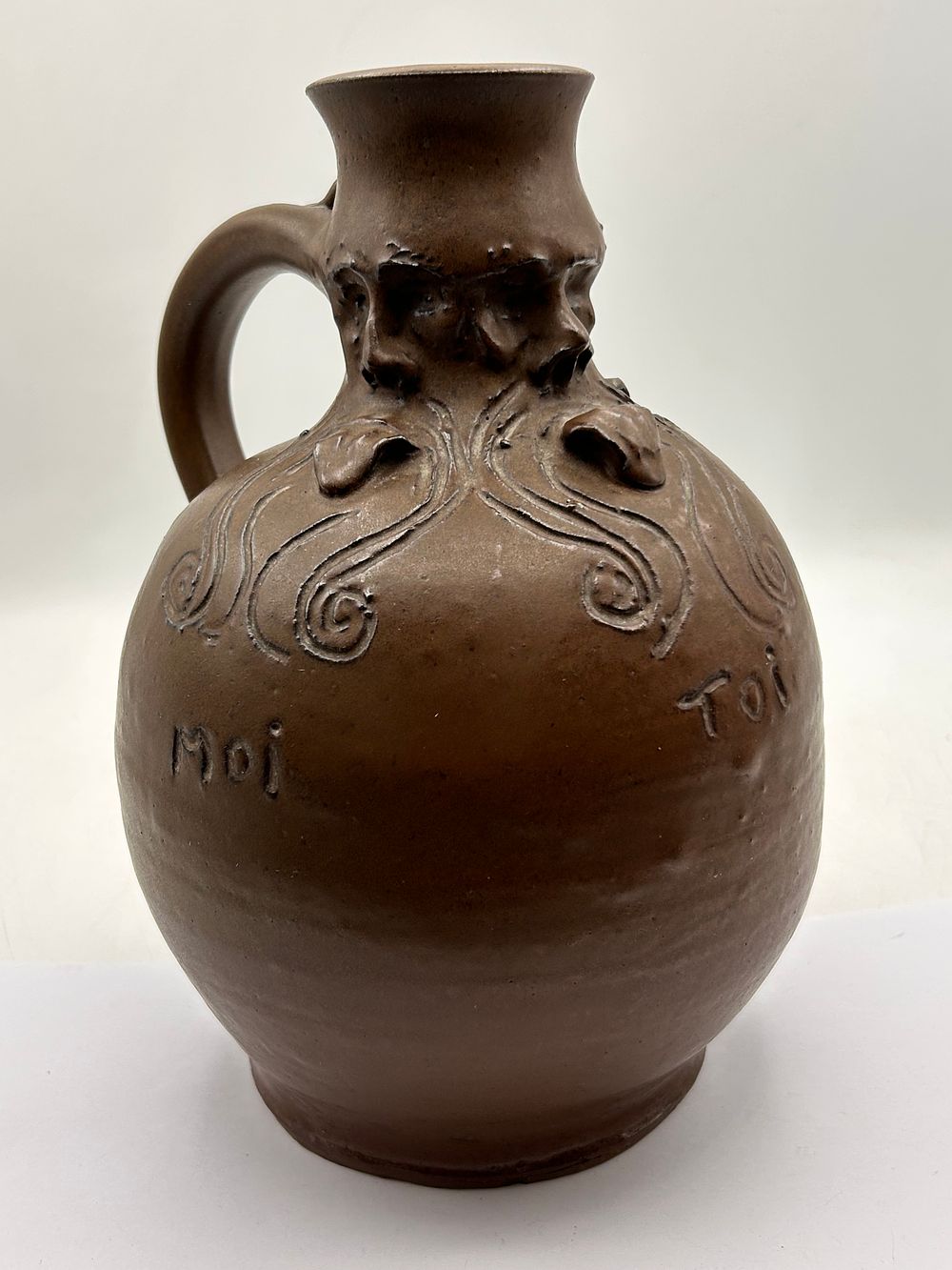 Lot 380: Dark Brown Stoneware Jug with Grotesque Faces and French Inscriptions, 9” Tall