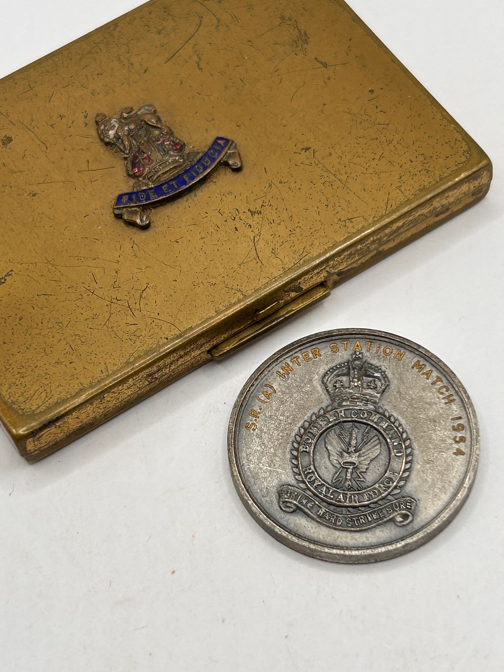 Lot 399: A Brass-Toned Military-Style Compact Case with 'FIDE ET FIDUCIA' Badge and a 1954 Royal Air Force Bomber Command Medallion