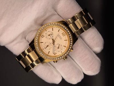 שעון Guess לאישה בצבע זהב עם קריסטלים שקופים בבזל החיצוני, לוח זהב פנינה ואינדקסים בצבע זהב. 