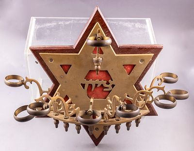 חנוכייה הודית לתלייה עשויה פליז ועץ. קנים מעוטרים בעבודה פתוחה, גב מעוטר במגן דוד וכיתוב 