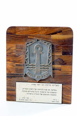 תבליט ברונזה עם הכיתוב "ופרצת ימה" מותקן על גב עץ זית מוקדש לעובד רשות הנמלים מצבי קינן יו"ר 