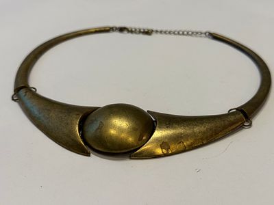 שרשרת קשיחה ודומיננטית בעיצוב אמנותי, עשויה מתכת בגוון ברונזה/פליז עם פטינה טבעית. במרכזה אלמנט 