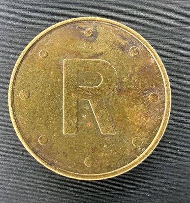 A brass token, with a large letter “R” surrounded by eight dots on one side and a decorative 