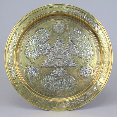 מגש פליז משובץ כסף ונחושת ומעוטר בקישוט צמחי וקליגרפיה באותיות ערביות . במרכז משולש גדול משובץ 