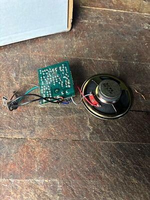 Electronic Circuit Board and Speaker Assembly. A green printed circuit board (PCB) and a small 
