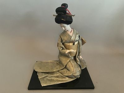 A vintage Japanese Geisha doll, depicted kneeling on a black rectangular base, with its head 