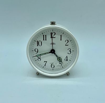 Prim alarm clock. A wind-up clock with a round body painted white, resting on three metal feet.